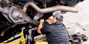 A close-up view of a car's exhaust system from underneath, showing the muffler, exhaust pipes, and catalytic converter with visible signs of wear and rust, set against the undercarriage of a lifted vehicle in a garage setting.